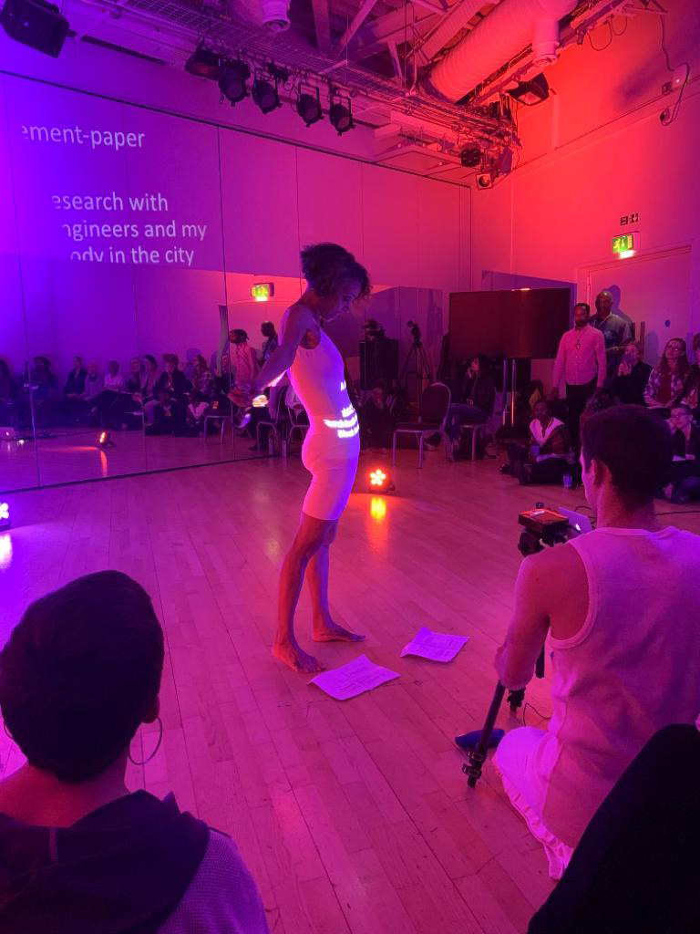 Dancer stand in middles of room lit in pink and purple light with text projected onto her body